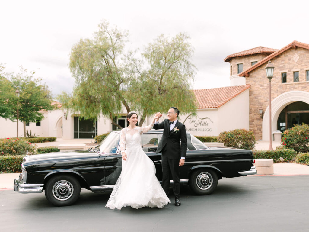 Bride and groom joyfully pose beside a classic black car in front of a Mediterranean-style building.