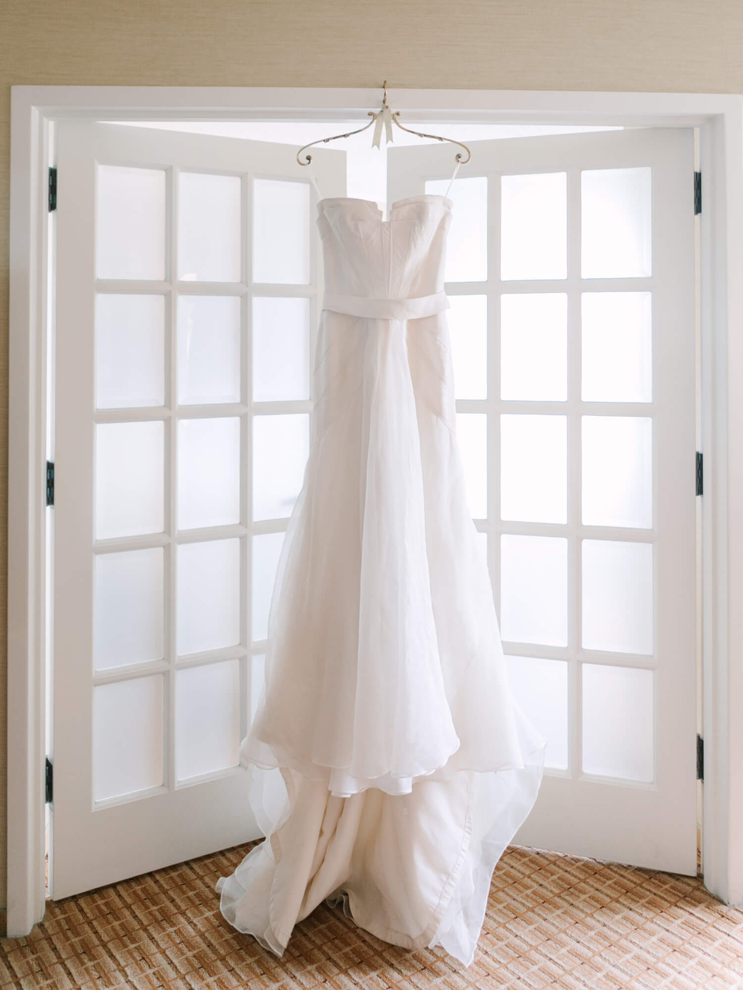 Strapless white wedding dress hanging on a hook against frosted glass French doors.