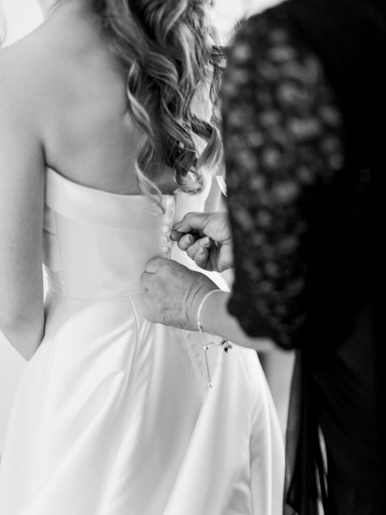 Black and white image of a bride in a white dress with long hair being buttoned by someone in lace.