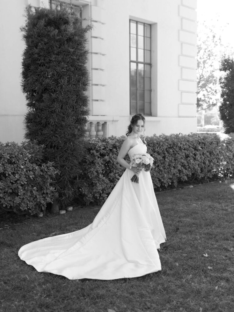 A bride in a flowing white gown stands serenely on a lawn. She holds a bouquet and is near a tall bush and elegant building.