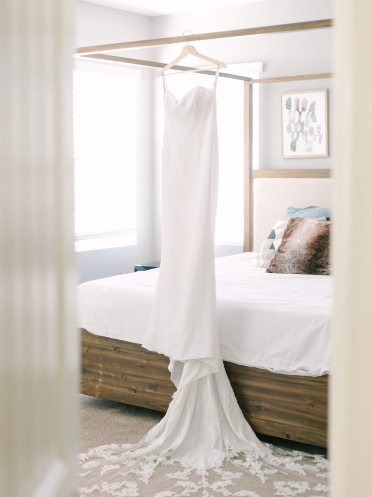 Elegant white wedding dress hangs on a wooden four-poster bed frame in a softly lit bedroom. The long train delicately drapes onto the carpet.