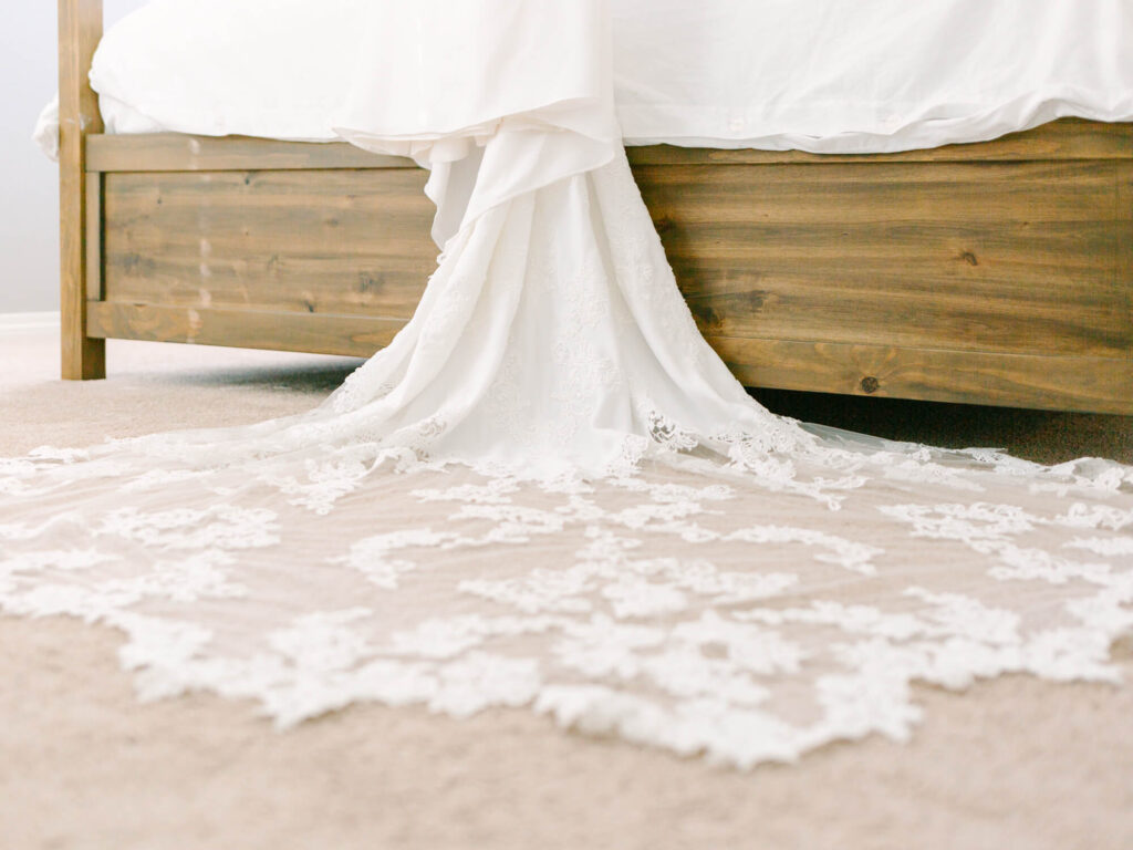 Long white lace wedding gown train elegantly spreads on beige carpet, flowing from a rustic wooden bed.