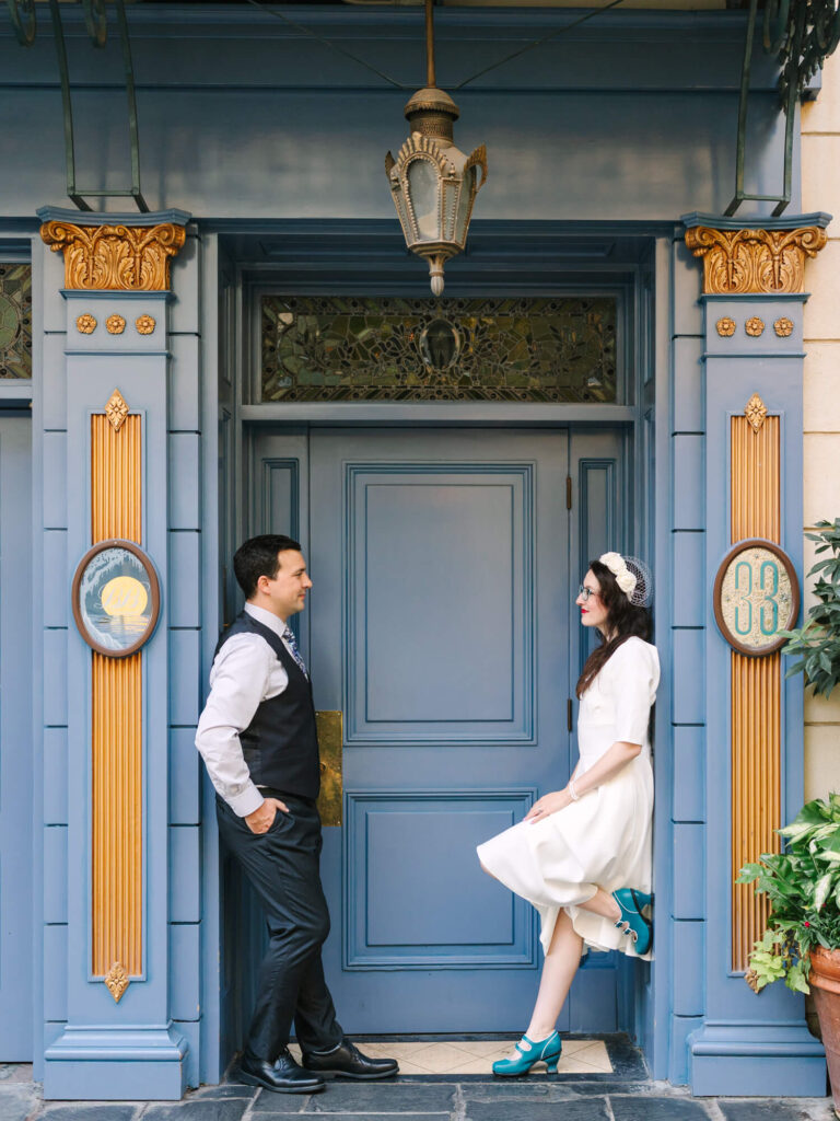 A couple smiles facing each other in front of an ornate blue door, with gold accents.