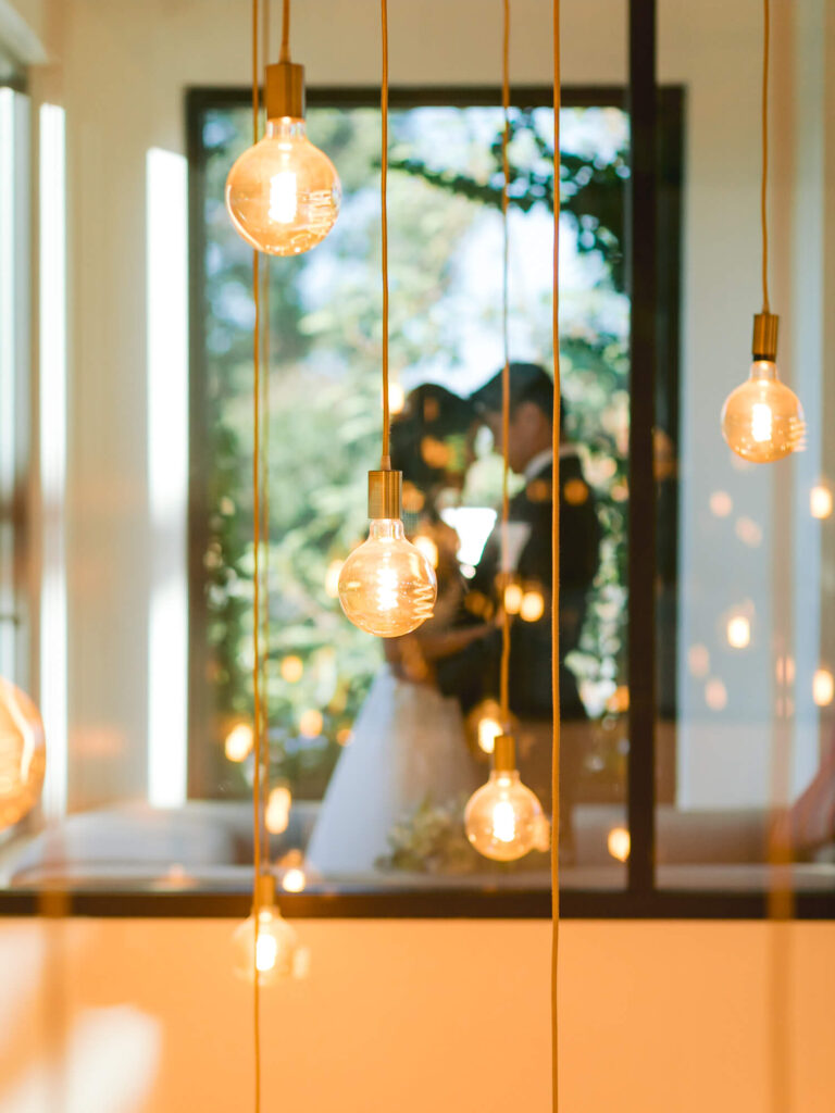 Softly lit hanging bulbs in focus, with a romantic silhouette of a couple embracing in the blurred background by a large window.