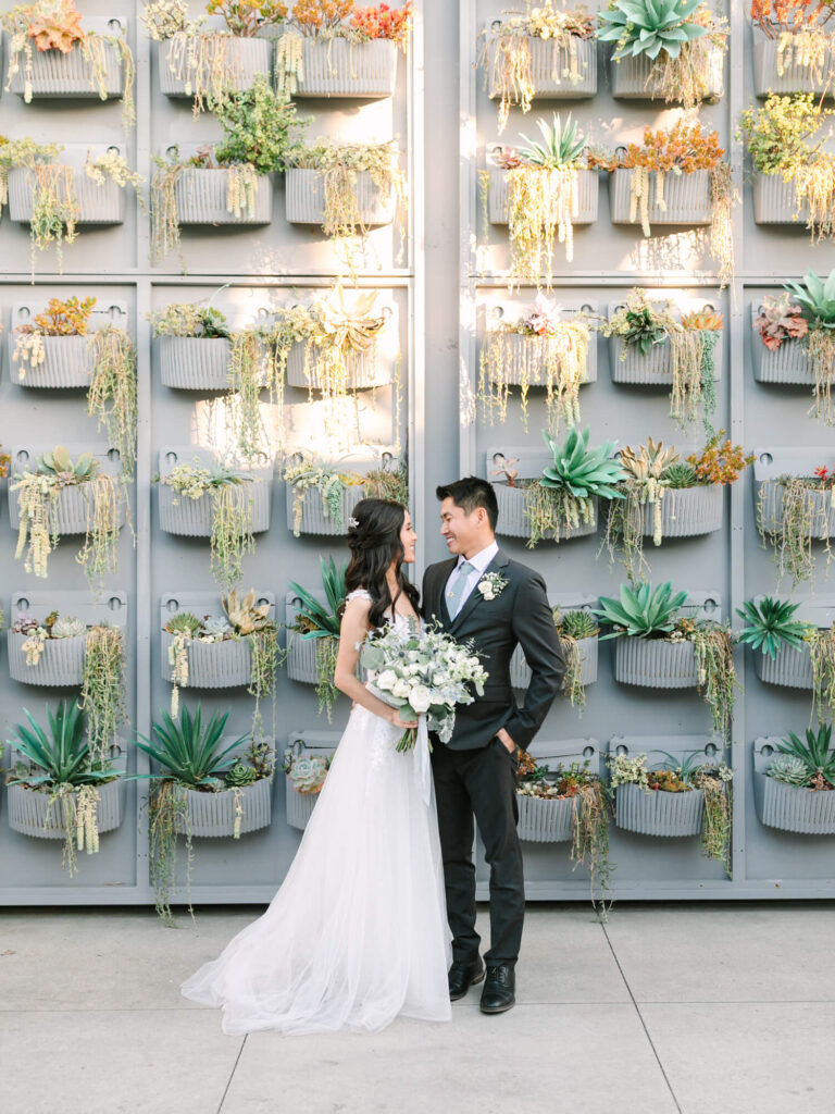 A bride in a white gown and groom in a dark suit stand smiling in front of a vertical succulent garden.