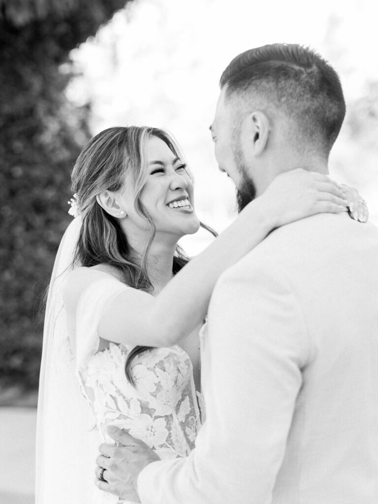 A joyful bride in a lace gown holds her partner's shoulders, smiling warmly at Coyote Hills.