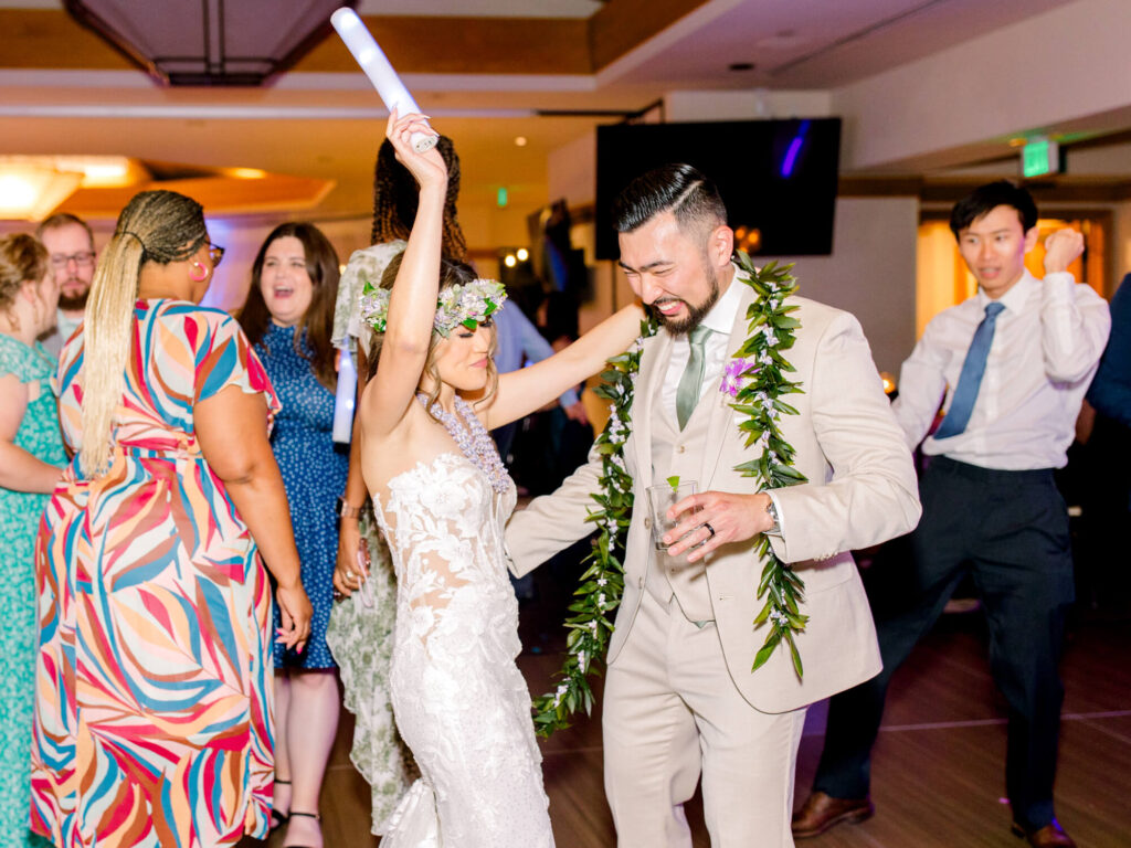 A joyful bride and groom dance closely at a lively wedding reception. The bride, in a floral crown, raises her hand, surrounded by smiling guests.