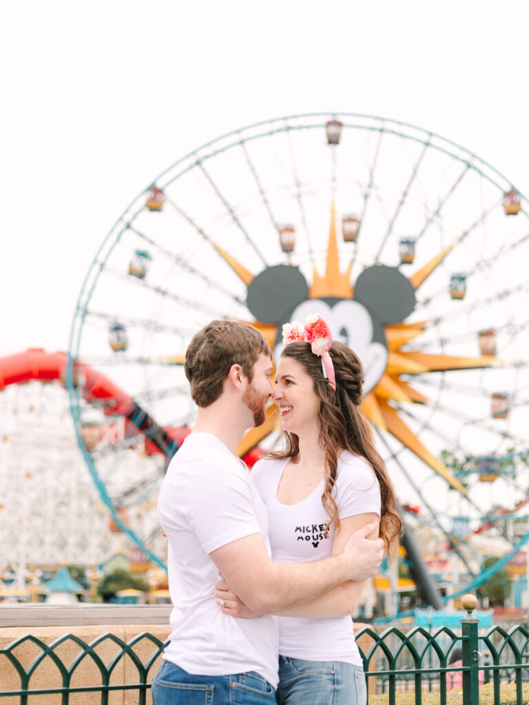 A couple embraces and smiles in front of a Ferris wheel featuring Mickey Mouse. Both wear white shirts, with the woman in Minnie Mouse ears.