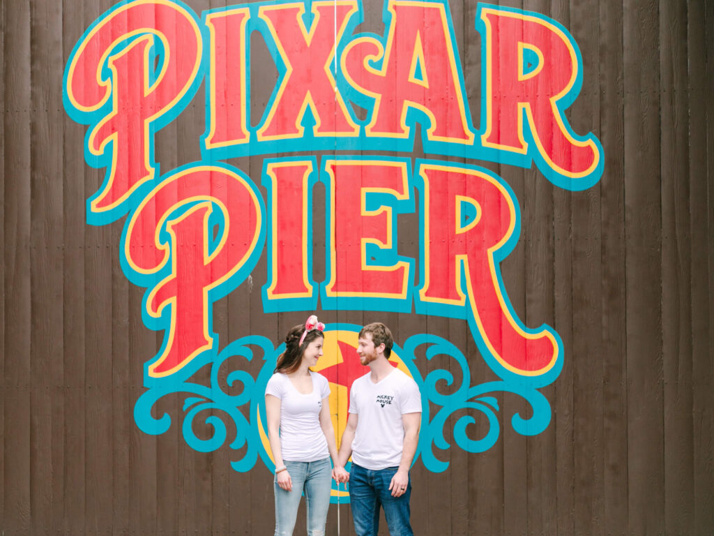 A couple stands smiling at each other in front of a vibrant "Pixar Pier" sign. They're wearing casual white T-shirts.