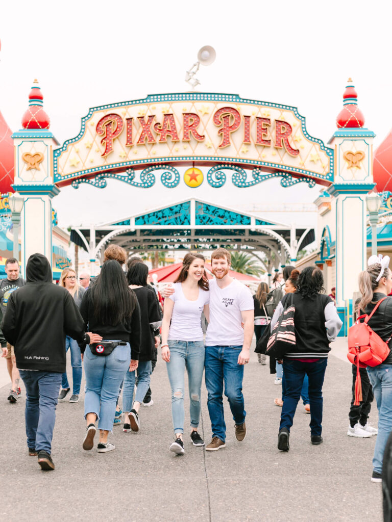 A smiling couple walks under a colorful "Pixar Pier" arch, surrounded by a lively crowd.