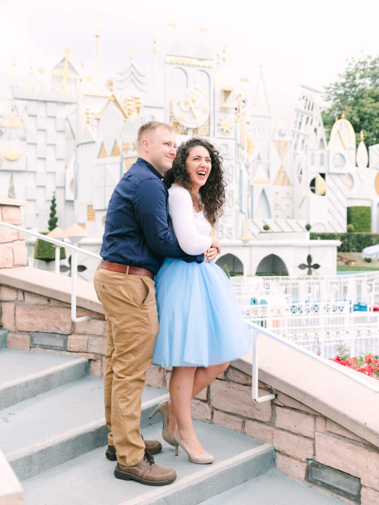 A couple embraces on stone steps, with a whimsical, castle-like structure in the background. The woman laughs.