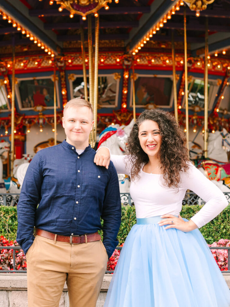 Two people smiling in front of a colorful carousel. The woman, in a flowing blue skirt, leans on the man’s shoulder.