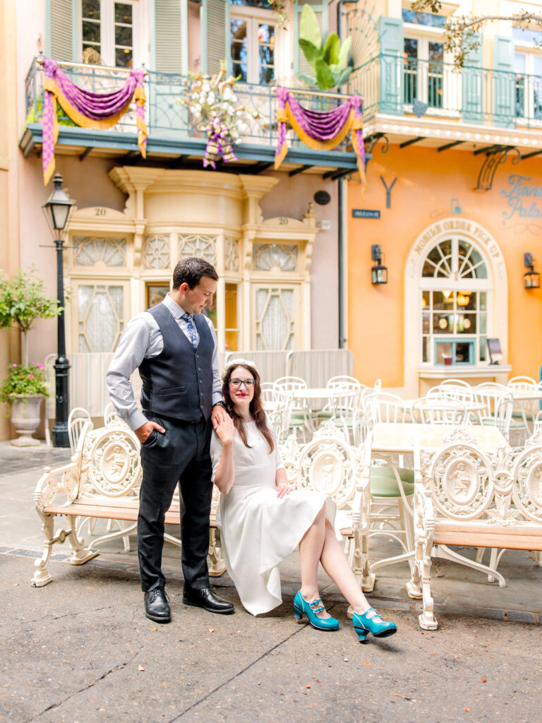 A couple poses in a charming courtyard with ornate white benches. She wears a white dress and teal heels, exuding elegance; he wears a vest and tie.