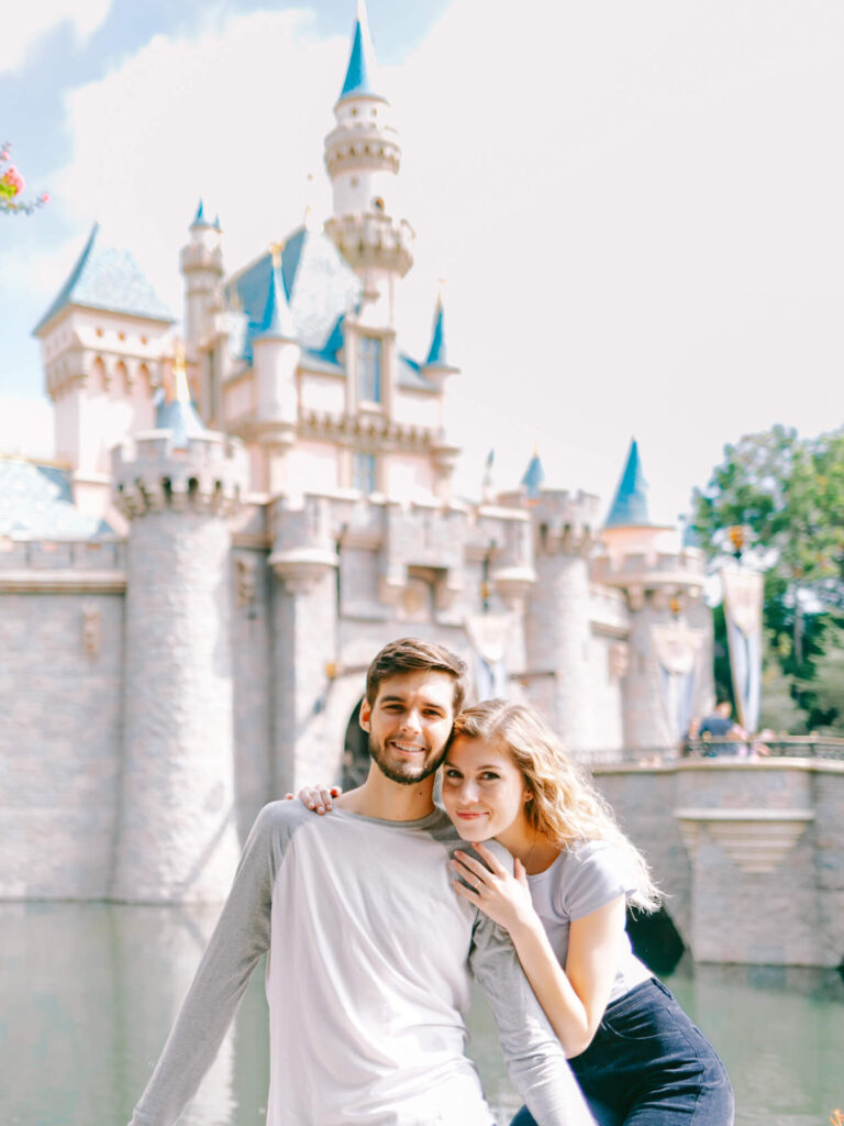 A smiling couple poses in front of a fairytale castle with blue spires, set against a bright sky.