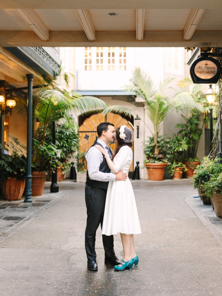 A couple embraces under an archway in a charming courtyard filled with potted plants and palm trees.