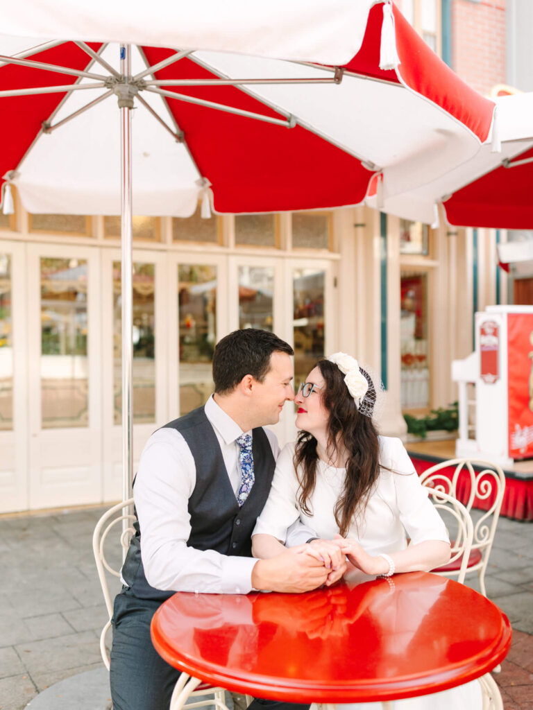 A couple sits at a red table under a red and white umbrella, gazing lovingly at each other.