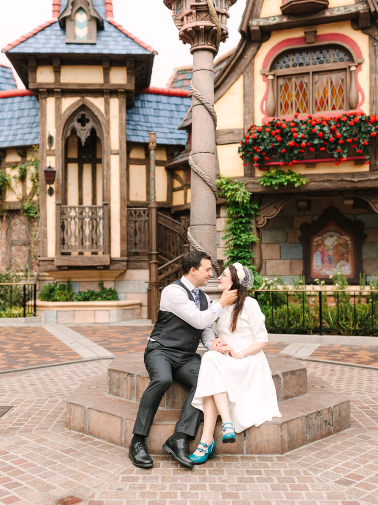 A couple sits affectionately on a stone bench in front of a charming, fairytale-like building adorned with flowers.