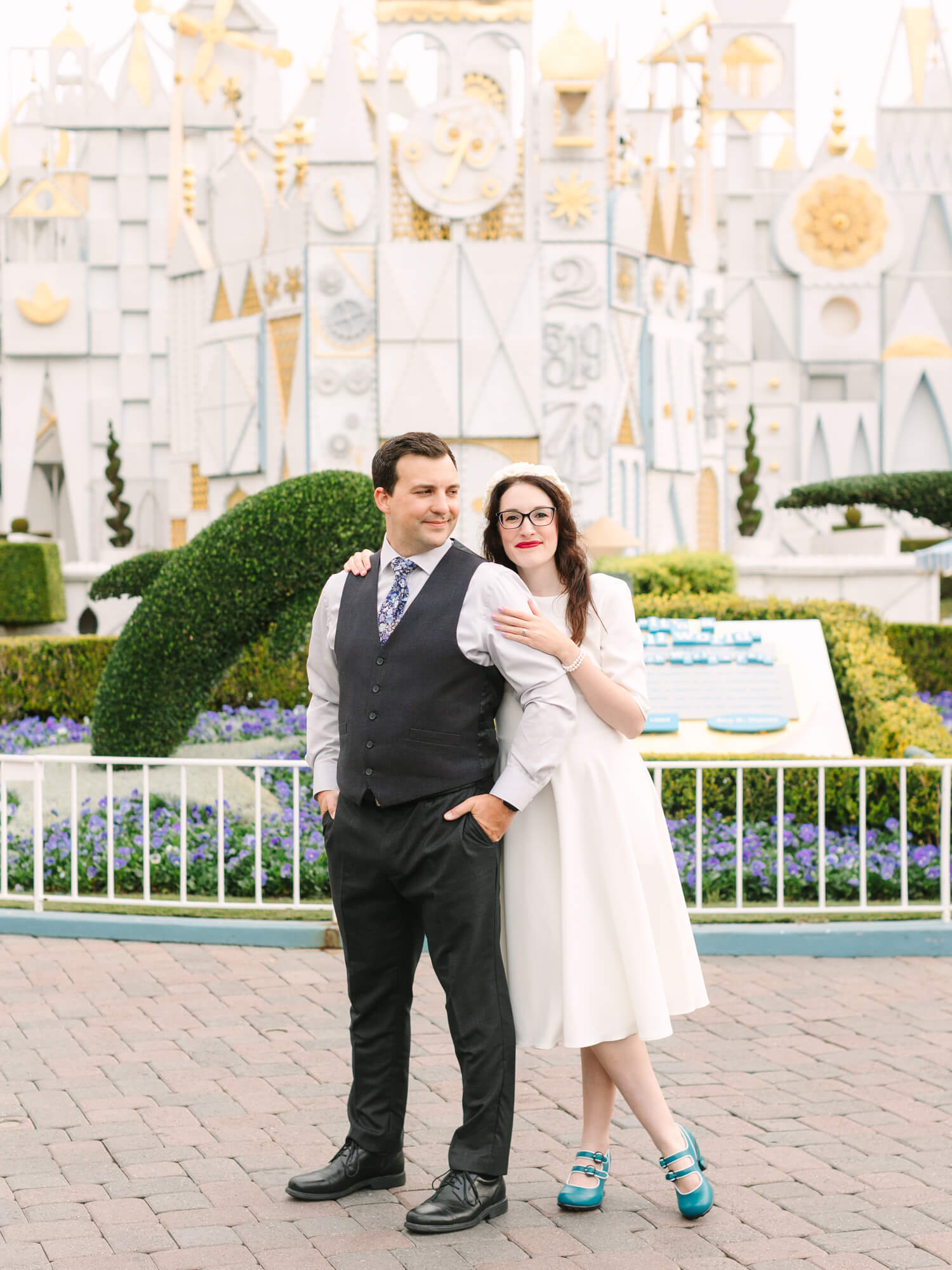 A couple poses in front of It's a Small World. The woman in a white dress lovingly holds the man's arm, who is wearing a vest and tie.