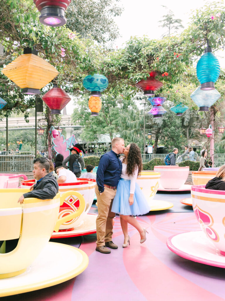 A couple shares a kiss in a colorful amusement park, standing among spinning teacup rides.
