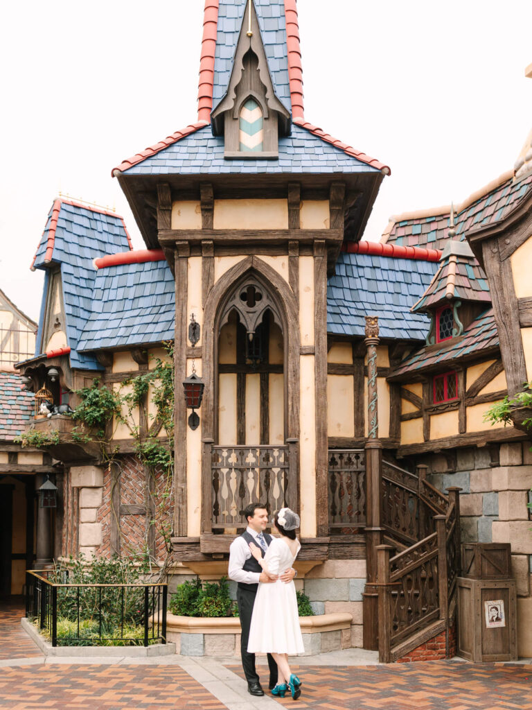 A couple embraces outside a whimsical, fairy-tale cottage with blue and red roofs, wooden accents, and arched windows.