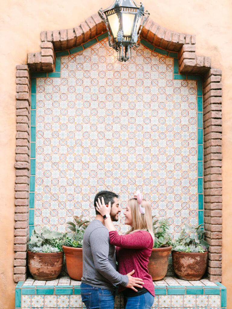 A couple stands close, embracing affectionately against a colorful tiled wall with potted plants beneath.