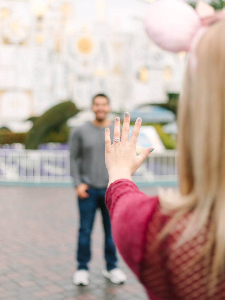 A woman in a red sweater shows an engagement ring while a man stands smiling in the blurred background.