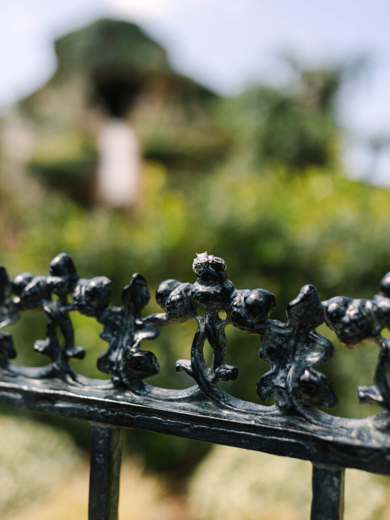 A small engagement ring rests on an ornate black metal fence with intricate designs.
