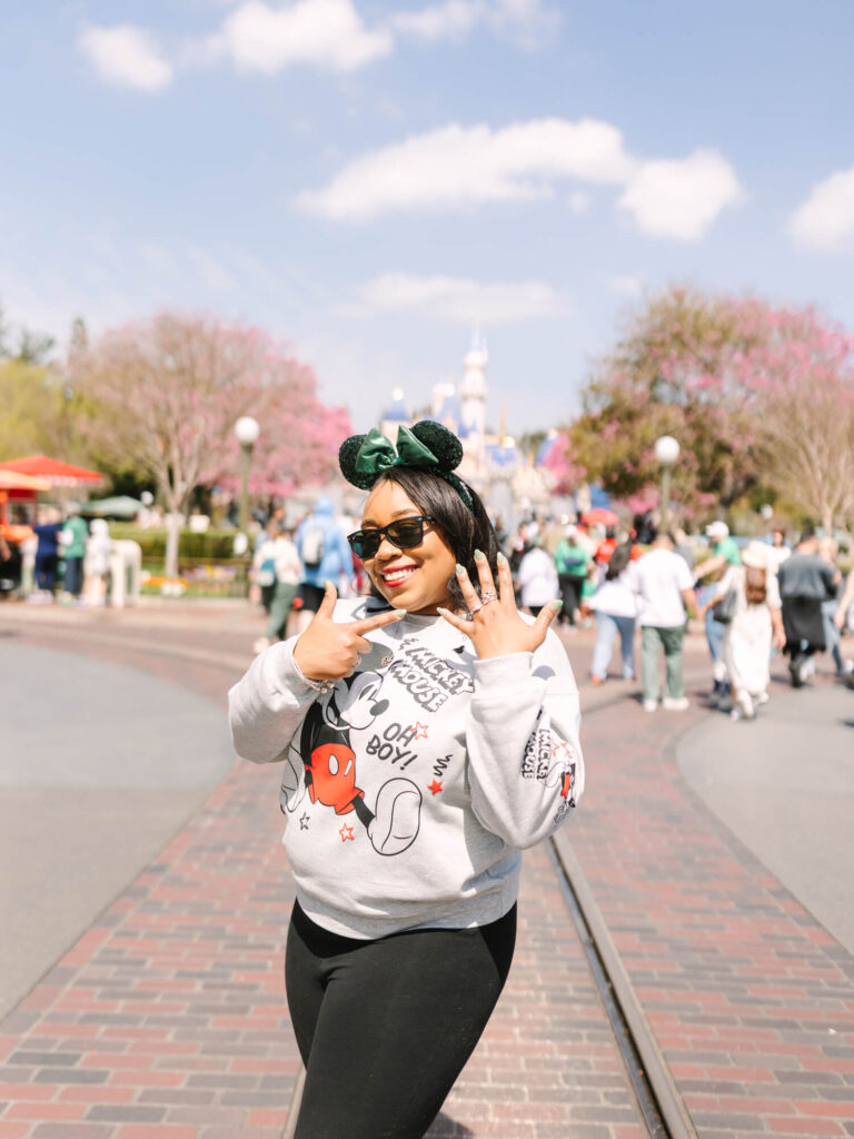 Woman wearing a Mickey Mouse sweater and Minnie Mouse ears poses happily in front of Disneyland theme park castle.
