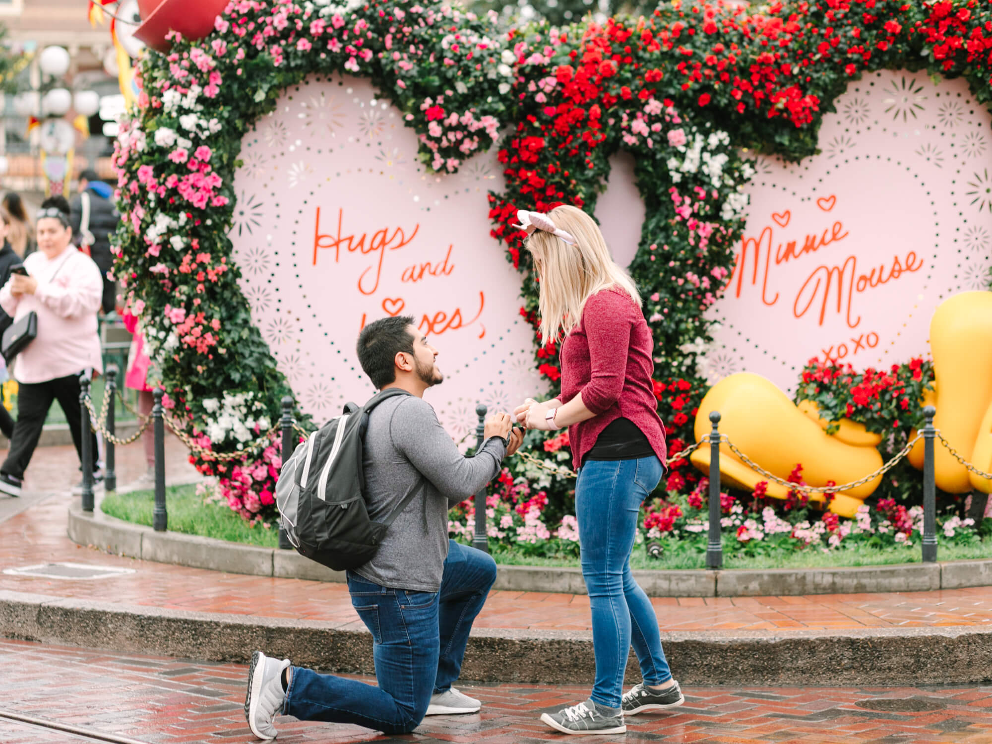 A man kneels in front of a woman in a romantic proposal scene at Disneyland. They are surrounded by vibrant heart-shaped floral displays.