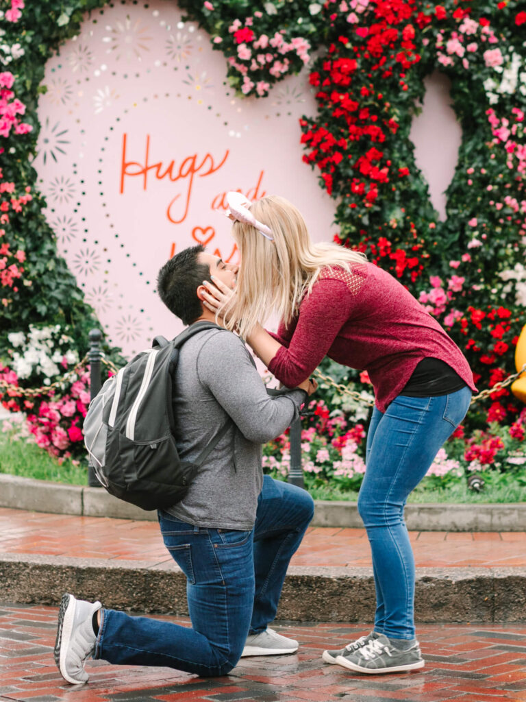A man kneels, proposing to a woman in front of a large heart-shaped floral backdrop with "Hugs and Kisses" written on it. She holds his face, smiling.