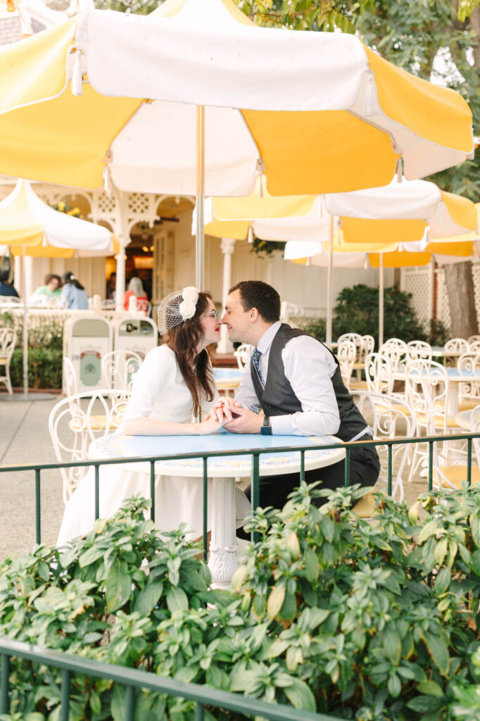 A couple leans over a table to kiss at an outdoor cafe with yellow-and-white umbrellas.