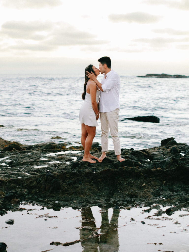 A couple embraces on rocky shore at sunset, wearing white clothes. Calm ocean in background, reflecting their silhouette in a water puddle nearby.