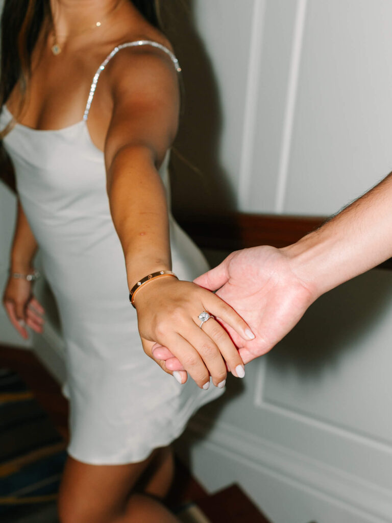 A woman in a white dress is holding hands with someone, showcasing a diamond ring.