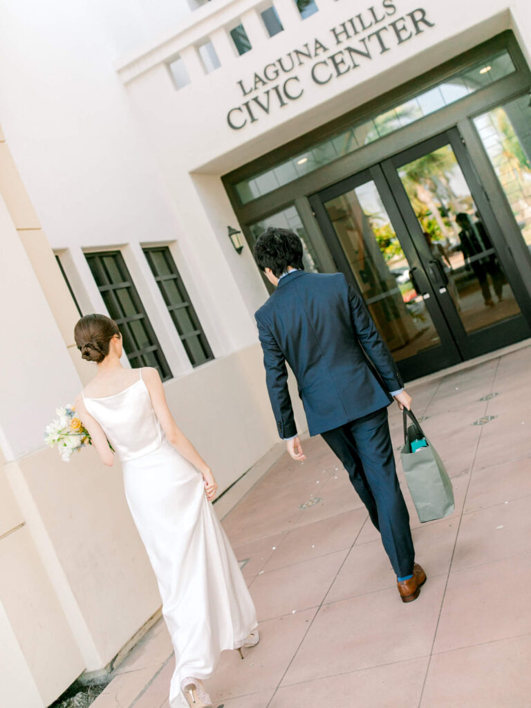 A couple walks towards double doors at Laguna Hills Civic Center. The woman wears a white dress and holds a bouquet; the man is in a suit, carrying a bag.