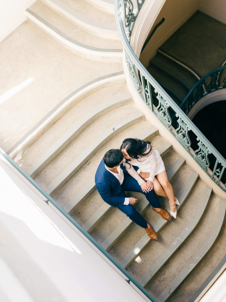 A couple sits intimately on curved stairs, viewed from above. The woman wears a white dress, the man a blue suit.