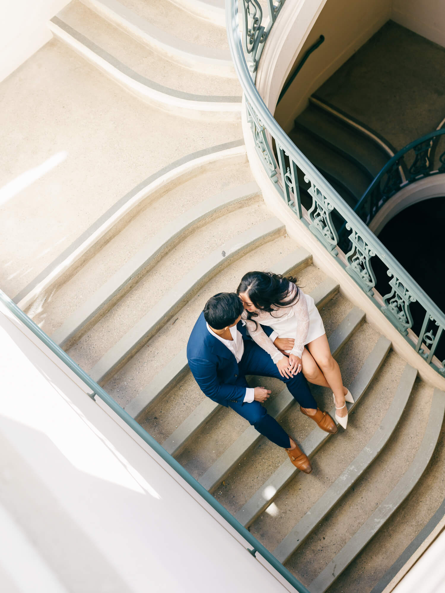 A couple sits intimately on curved stairs, viewed from above. The woman wears a white dress, the man a blue suit.