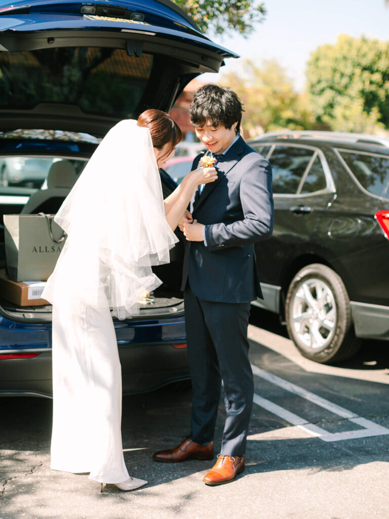 Bride in a veil adjusts groom's boutonniere near an open car trunk.