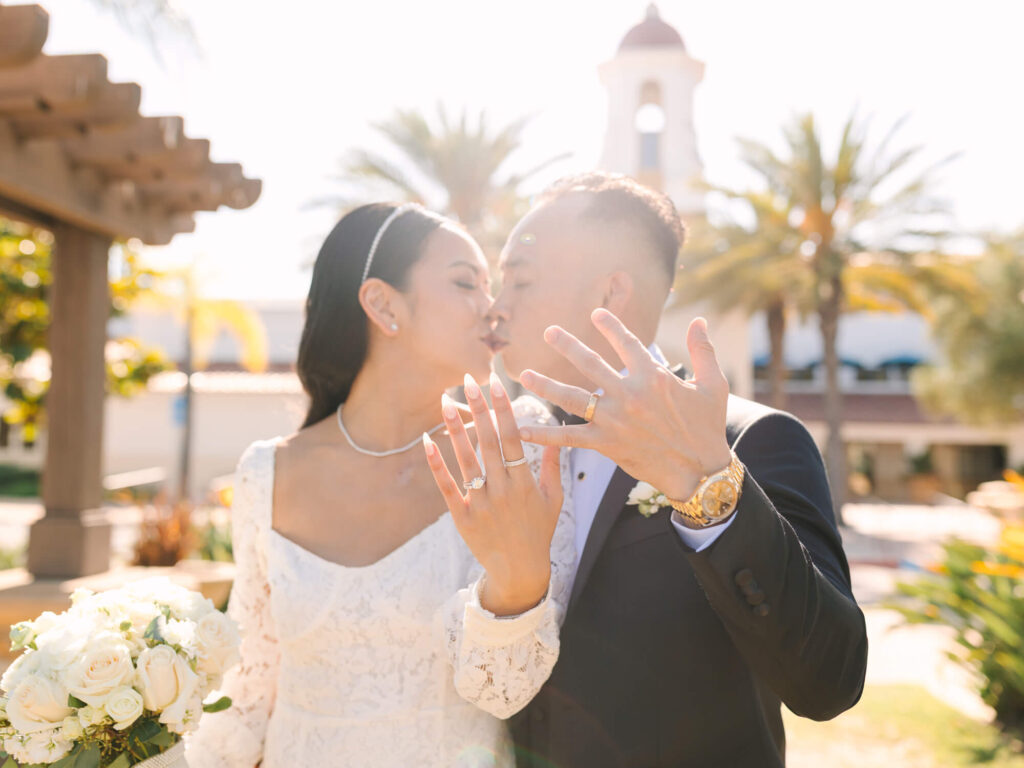 Bride and groom share a kiss outdoors, showcasing their wedding rings. The bride holds a bouquet of white roses.