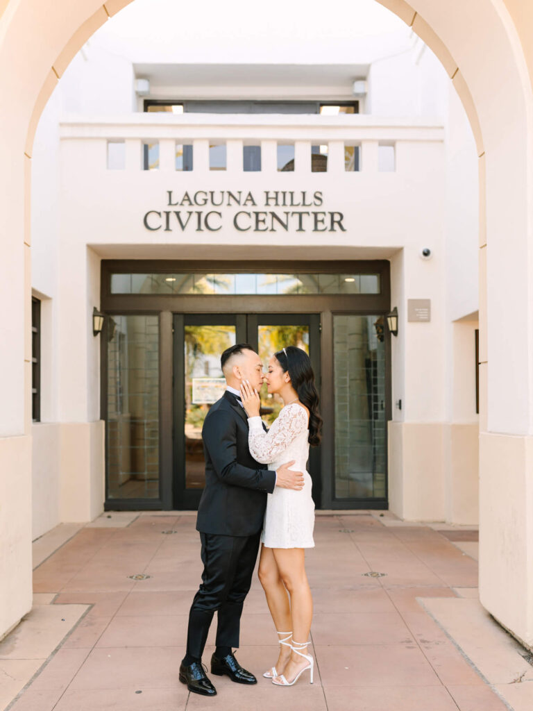 A couple embraces lovingly in front of Laguna Hills Civic Center. The man wears a suit, and the woman in a white dress smiles.