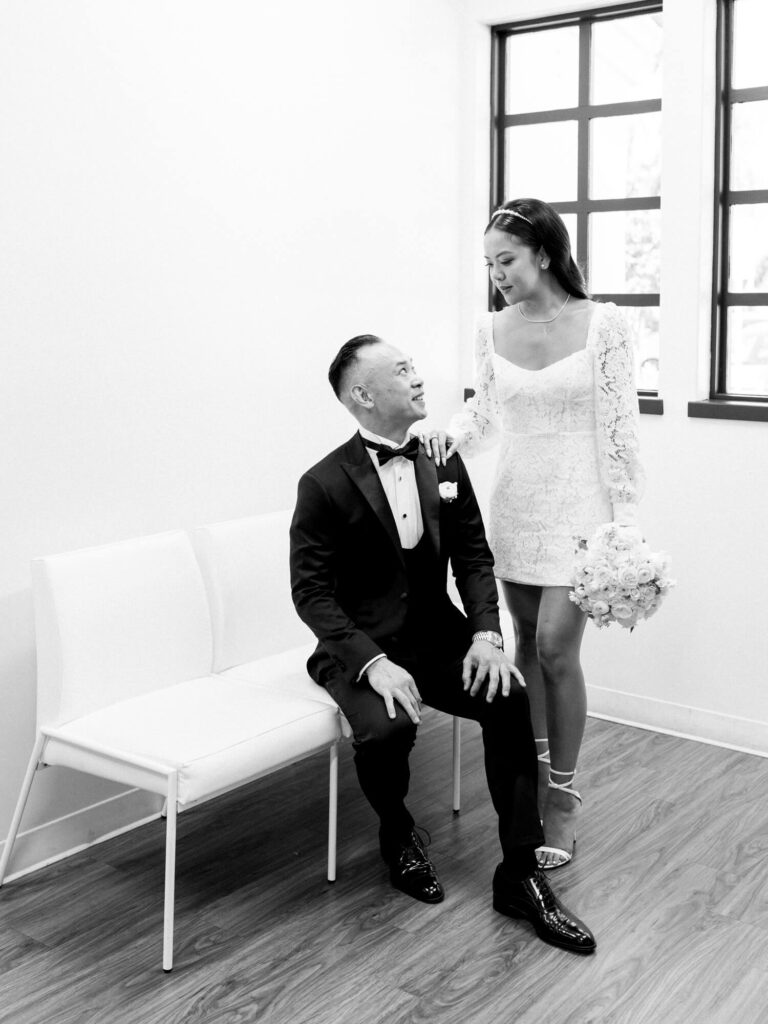 Black and white photo of a bride holding flowers, stands beside the groom seated on a bench, both smiling warmly.