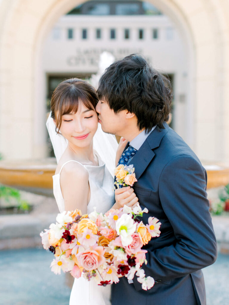 A groom in a blue suit kisses the bride's cheek, as she smiles softly, holding a vibrant bouquet.