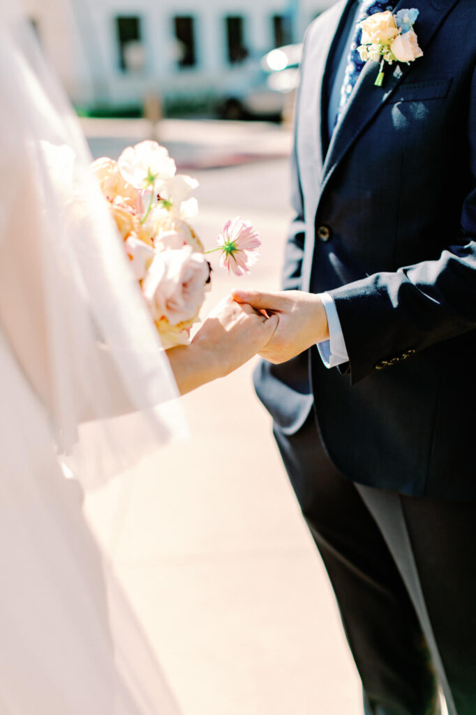 A groom in a dark suit gently holds hands with a bride in a white dress. The bride holds a bouquet of light pink flowers.