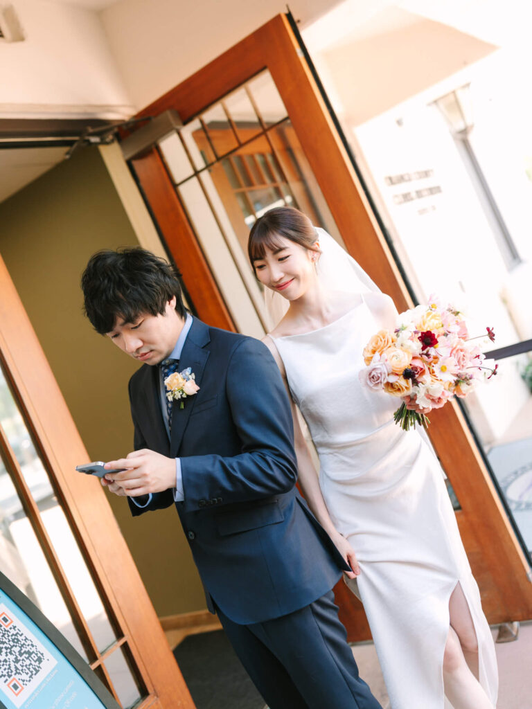 A bride in a white dress with a bouquet smiles warmly at a groom in a navy suit, who is looking on his phone
