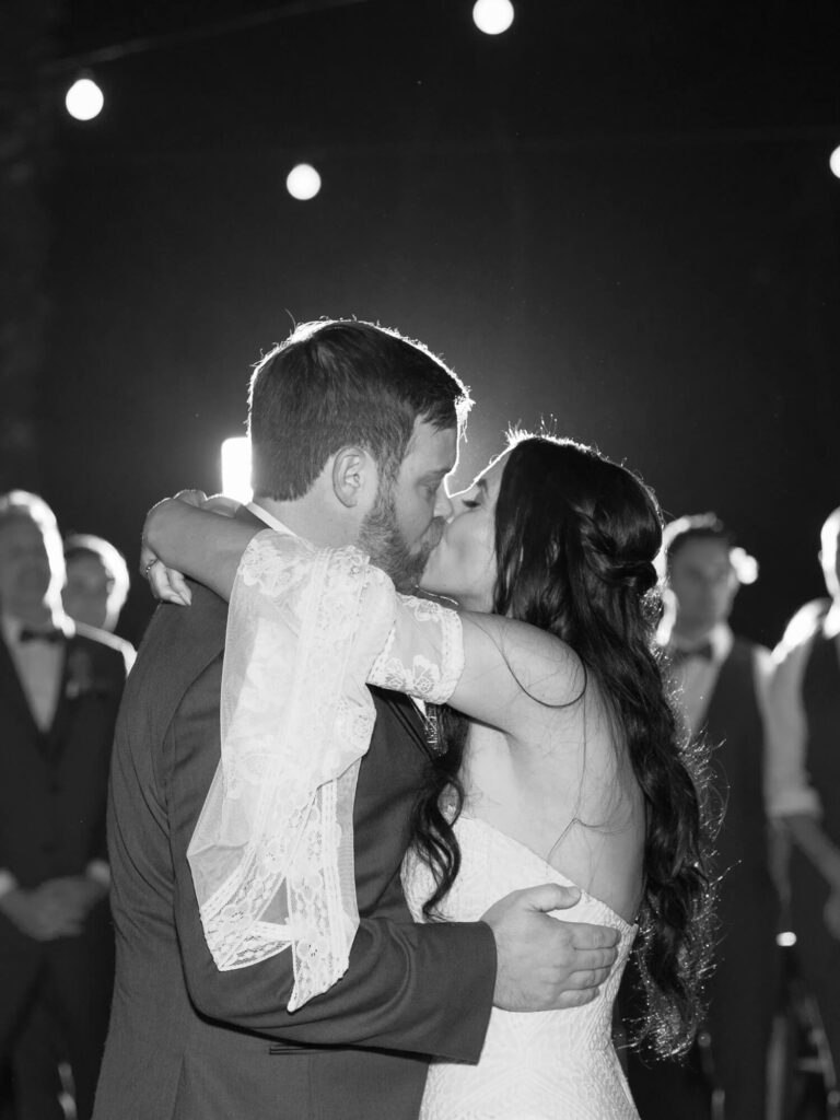 A bride and groom share a kiss on the dance floor at night, surrounded by blurred onlookers.