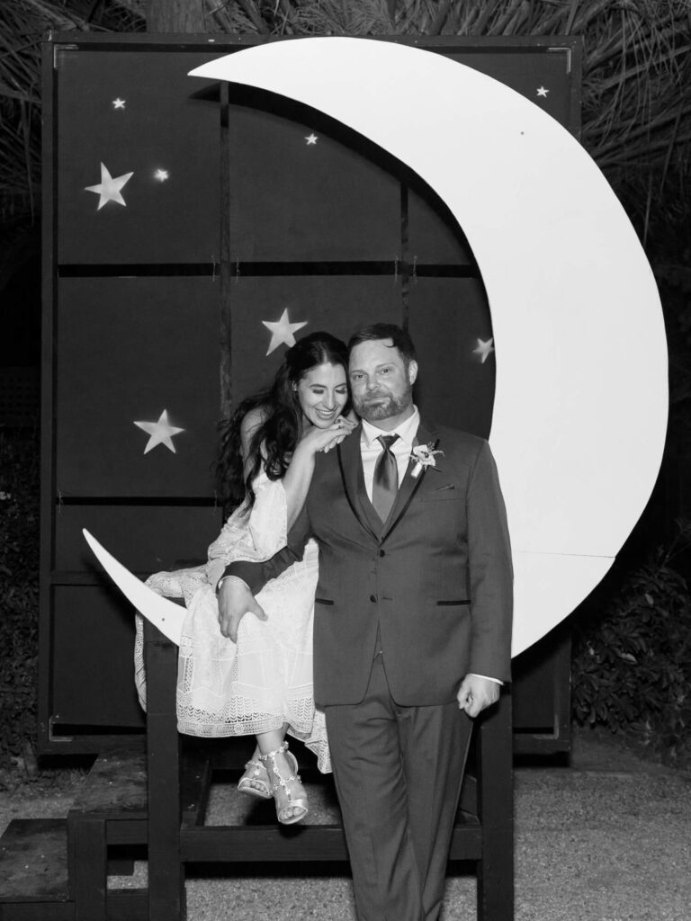 A joyful couple poses in front of a large crescent moon and star backdrop. The woman sits on the moon, smiling and leaning on the man's shoulder.