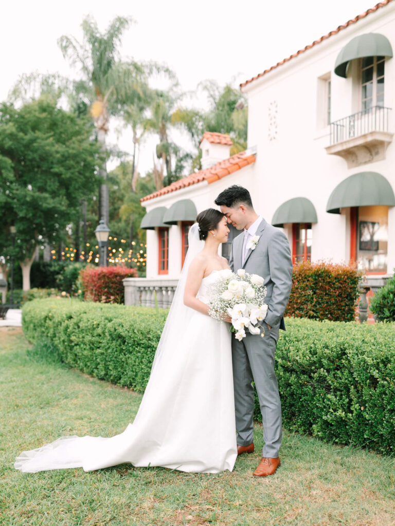 A bride in a flowing white gown and groom in a gray suit stand closely, sharing an intimate moment.