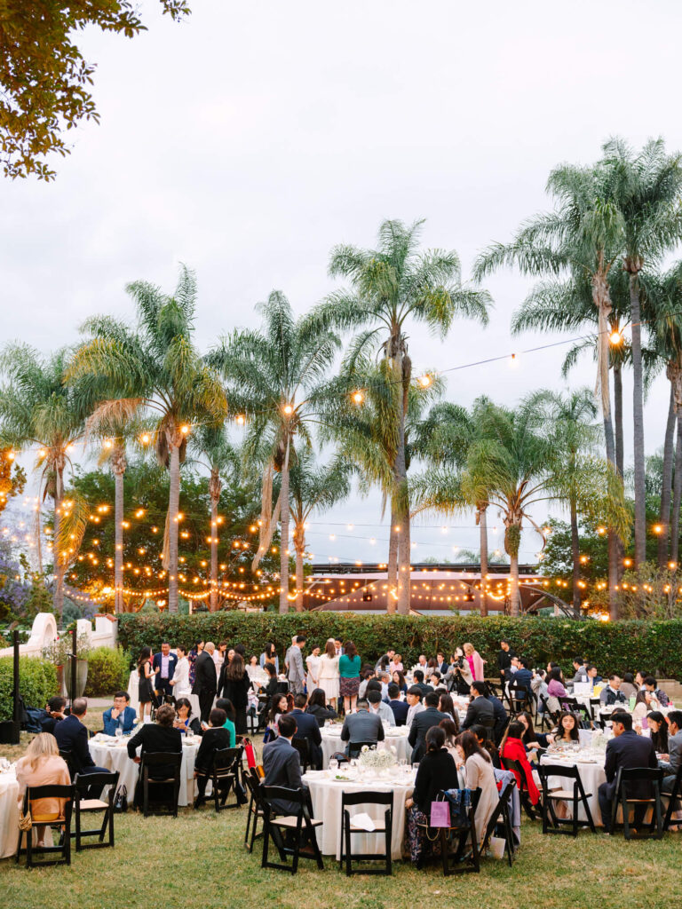 Outdoor wedding reception under palm trees with string lights at dusk. Guests in formal attire sit at round tables.