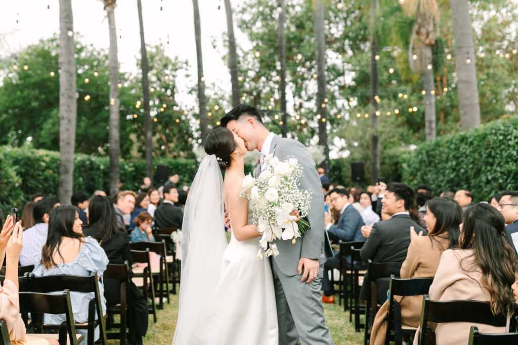 A bride and groom kiss outdoors under string lights, surrounded by seated guests.