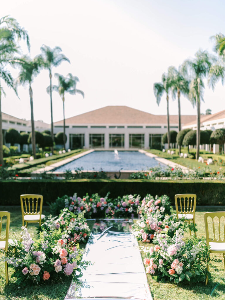 Elegant garden wedding setup with a mirrored aisle, surrounded by colorful flowers and gold chairs.