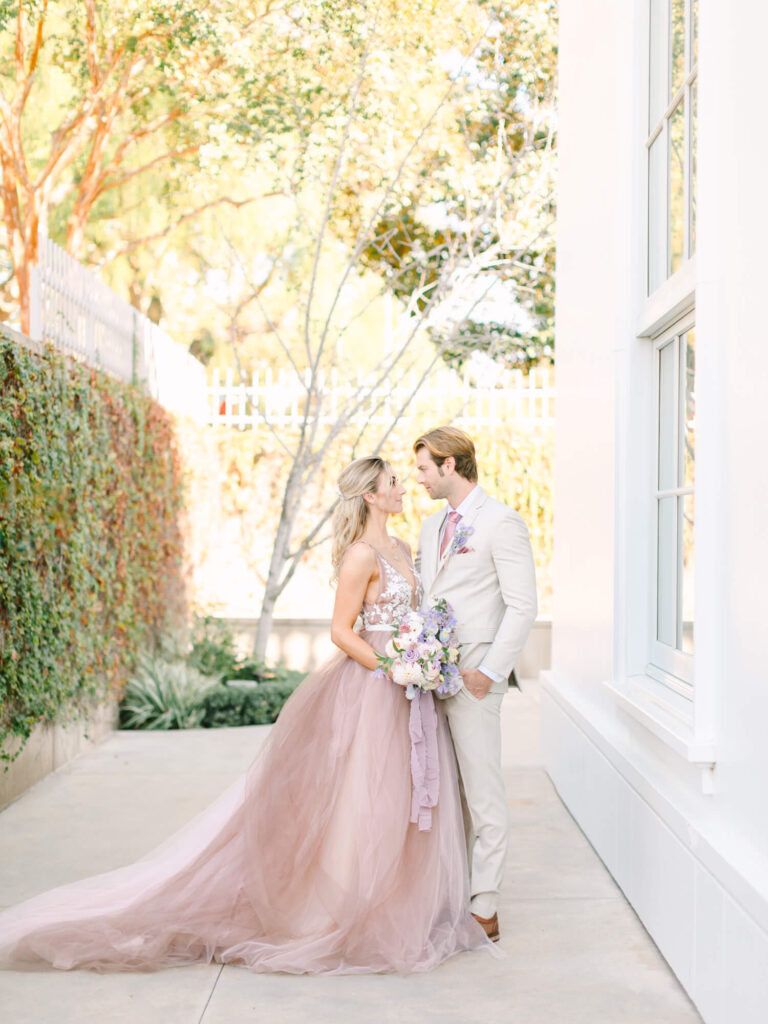 A couple embraces outdoors, surrounded by greenery. The bride wears a flowing pink gown and holds a bouquet.